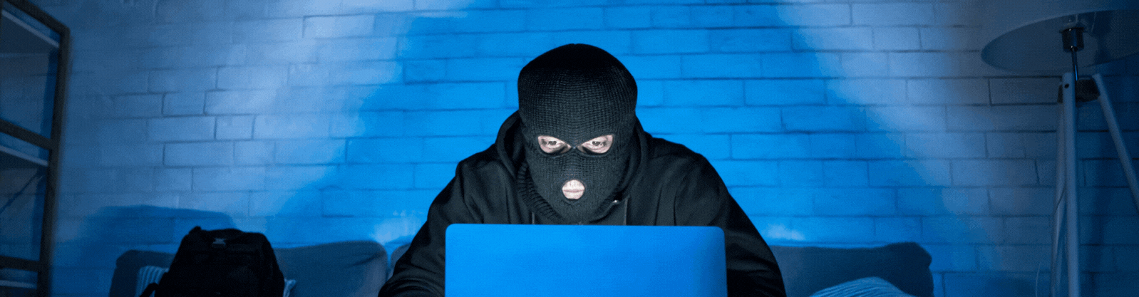 Ein Mann in schwarzer Kleidung und schwarzer Sturmhaube, der vor einem Notebook sitzt. Er repräsentiert einen Hacker, der als Beispiel für Cybercrime und Gefahren im Internet und Netzwerken steht. Das Bild dient als Beitragsbild des IT Security Awareness Training.
